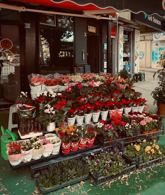 An outdoor display of fresh flowers in various arrangements and colors, including pink, red, white, and purple blossoms, positioned on black plastic and wooden crates outside a flower shop. The flower