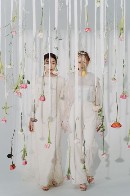 Two women dressed in elegant, long white dresses stand behind sheer white curtains decorated with hanging pink, red, and purple flowers, creating a delicate floral backdrop. The women have natural mak