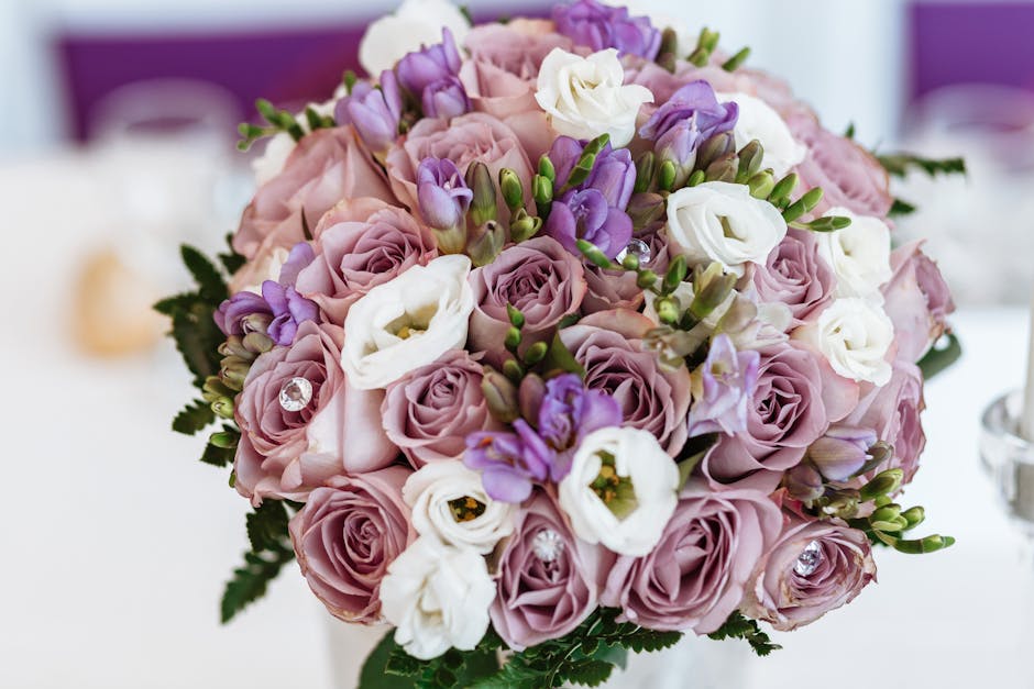 A close-up view of a floral bouquet featuring light pink roses, white lisianthus, and purple freesia arranged in a rounded, compact style. The flowers are fresh with smooth petals and vibrant colors,