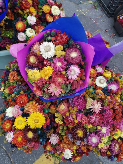 A vibrant bouquet of small, colorful flowers including daisies, chrysanthemums, and carnations in shades of pink, yellow, orange, white, and purple, wrapped in purple and pink tissue paper. The bouque